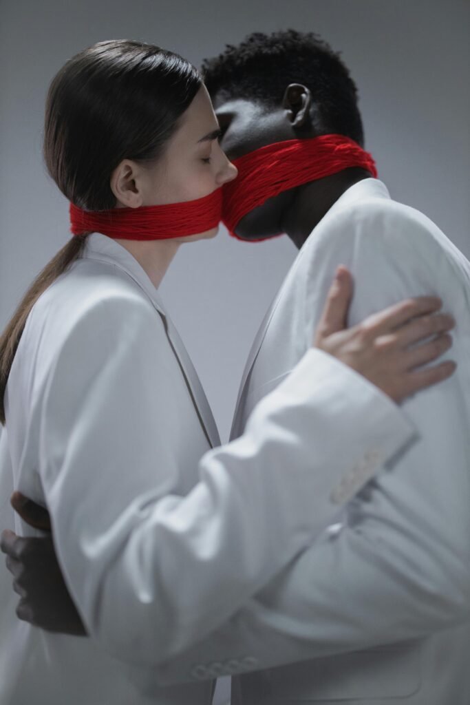 Home Artistic portrayal of a couple embracing with red fabric covering their mouths, symbolizing silence.